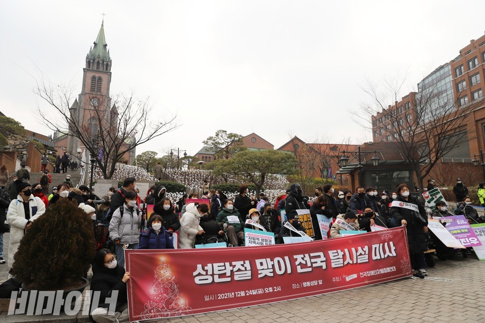 미사 현장. 빨간 현수막에 트리가 그려져 있고 ‘성탄절 맞이 전국 탈시설 미사’라고 적혀 있다. 활동가들 뒤로 거대한 명동성당이 보인다. 사진 하민지