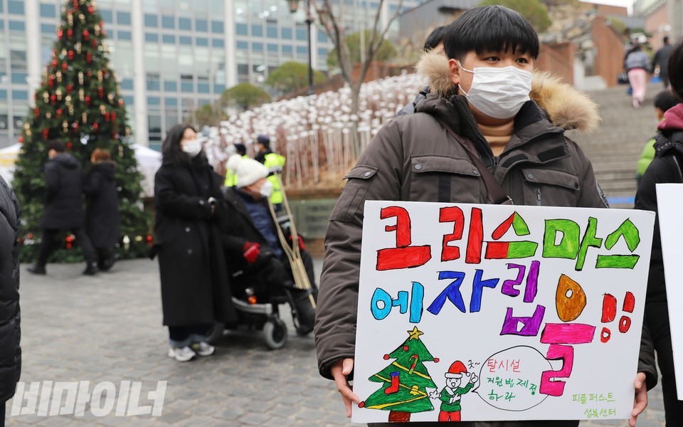 피플퍼스트성북센터 활동가가 ‘크리스마스에 자립을’이라 적은 피켓을 들고 있다. 피켓 속 그림에 산타가 ‘탈시설지원법 제정하라’라고 외치고 있다. 왼쪽에 커다란 트리가 보인다. 사진 하민지