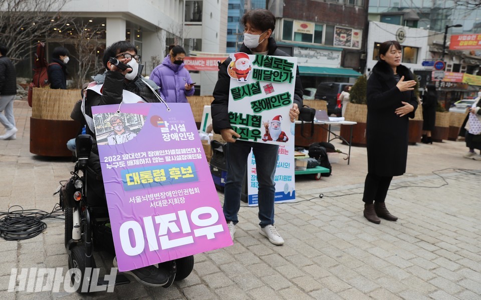 이진우 대선후보가 발언하고 있다. 오른쪽에는 이진우 후보를 지지하는 활동가가 ‘시설로는 배달 안 가, 탈시설장애인당, 한뇌협 이진우’라고 적힌 피켓을 들고 웃으며 이 후보를 바라보고 있다. 수어통역을 하는 통역사도 보인다. 사진 하민지