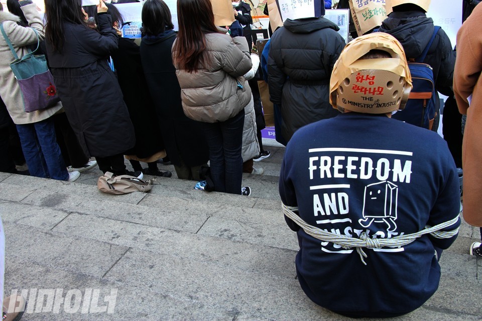 화성외국인보호소 고문 피해자 M 씨의 뒷모습. 머리 보호대를 착용하고 몸이 밧줄에 묶여진 채 기자회견장에 앉아 있다. 보호소에서 당한 고문을 재현한 것이다. M 씨가 입은 티셔츠에는 영어로 ‘프리덤 앤 저스티스(자유와 정의)’라고 적혀 있다. 머리 보호대에는 ‘화성 관타나모’라고 적혀 있다. 사진 하민지