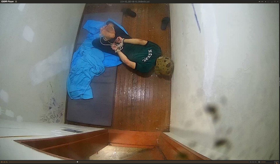 화성외국인보호소 내 CCTV 화면. ㄱ 씨가 일명 '새우꺾기 고문'을 당한 채 독방에 격리돼 있다. 묶인 손목과 발목이 등 뒤에서 연결돼 있고 ㄱ 씨의 배가 바닥에 닿아 있다. ㄱ 씨의 머리에는 보호대가 씌워져 있으며, 상의에는 ‘보호외국인’이라 적혀 있다. 방이 매우 좁아 보인다. 사진 사단법인 두루