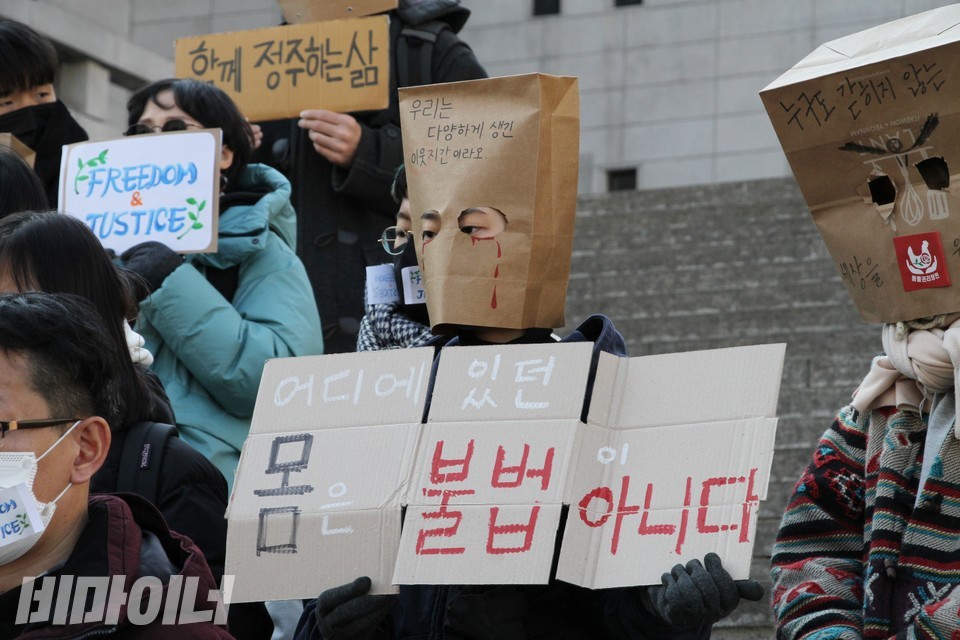 한 활동가가 기자회견장에 봉투가면을 쓰고 참석했다. 봉투가면에는 “우리는 다양하게 생긴 이웃지간이라오”라고 적혀 있다. 가면은 눈 부분만 뚫려 있는데, 피눈물을 흘리는 그림이 그려져 있다. 활동가가 든 피켓에는 “어디에 있던 몸은 불법이 아니다”라고 적혀 있다. 사진 하민지