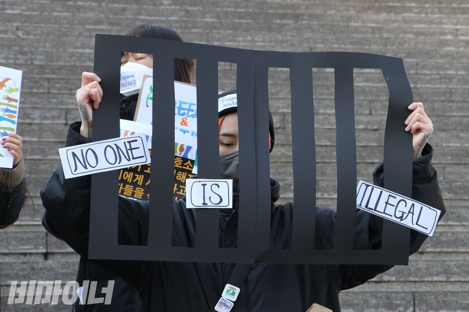 한 활동가가 있다. 검은색 쇠창살을 재현한 피켓을 들고 있다. 피켓에는 “불법으로 존재하는 사람은 없다(No one is illegal)”고 젹혀 있다. 사진 하민지