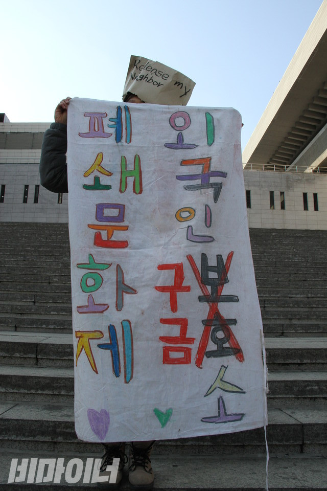 한 활동가가 하얀 천에 “외국인구금소 폐쇄 문화제”라 적은 피켓을 들고 서 있다. ’보호’에 X표시를 치고 ’구금’이라 적었다. 그가 쓴 봉투가면에는 ’내 이웃을 풀어줘라(Release my neighbor)’라고 적었다. 사진 하민지