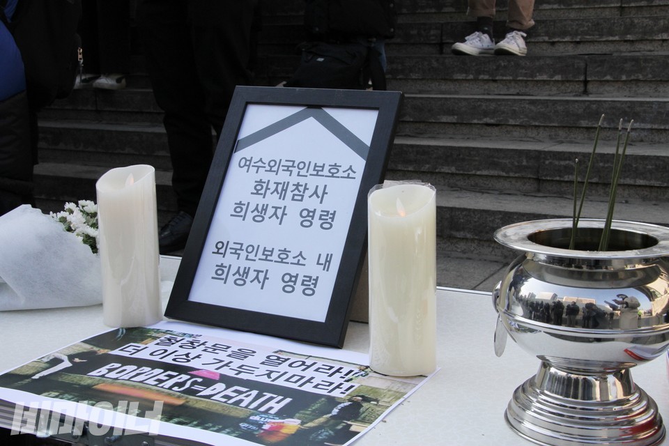 여수외국인보호소 화재참사 희생자 추모 테이블. 희생자 영령을 표현한 액자, 하얀 초, 향, 국화꽃이 있다. “철창문을 열어라! 더는 가두지 마라!”라고 적힌 피켓도 보인다. 사진 하민지