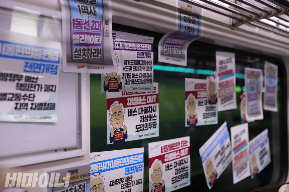 지하철 안 창문에 피켓 여러 장이 붙어 있다. 이재명 대선후보의 그림이 그려져 있고 ‘1역사 1동선 2대 엘리베이터 설치하라’, ‘차별버스 OUT, 버스 대폐차시 저상버스 도입 의무화’, ‘교통약자이동편의증진조례 전면 개정, 경기도 특별교통수단 수도권 전체 지역 운행’ 등이 적혀 있다. 사진 하민지