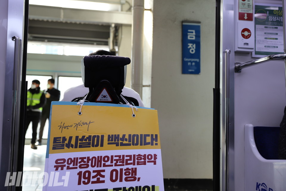 한 활동가가 금정역에서 열차 출입문을 막고 있다. 그는 휠체어에 ‘탈시설이 백신이다! 유엔장애인권리협약 19조 이행’이라 적힌 피켓을 걸었다. 사진 하민지