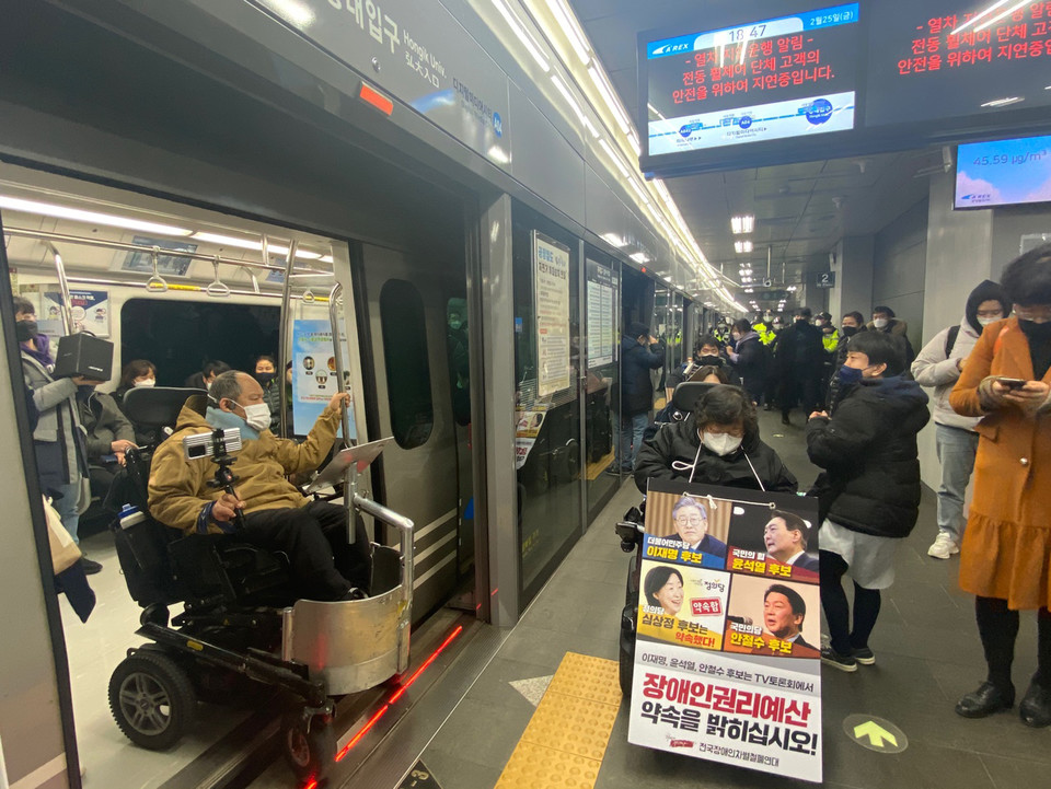 전국장애인차별철폐연대가 25일, 4차 TV 토론회를 앞두고 대선후보들에게 장애인권리예산 보장 약속을 촉구하는 지하철 연착 투쟁하고 있다. 상단에 있는 지하철 방송 화면에는 “열차 지연 운행 알림. 전동휠체어 단체 고객의 안전을 위하여 지연 중입니다”라는 문구가 송출되고 있다. 사진 전국장애인차별철폐연대