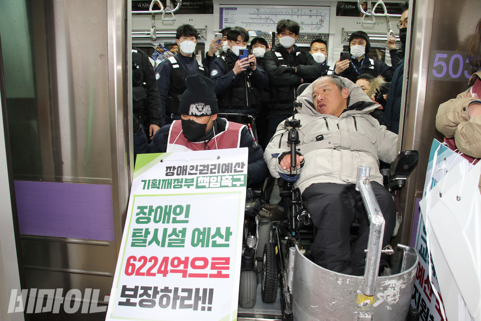 여의도역에서 휠체어 탄 장애인들이 지하철 문을 막아서고 있다. 그 뒤로 검은 옷을 입은 서울교통공사 지하철 보안관들이 핸드폰으로 촬영을 하거나 팔짱을 낀 채 이를 바라보고 있다. 사진 강혜민