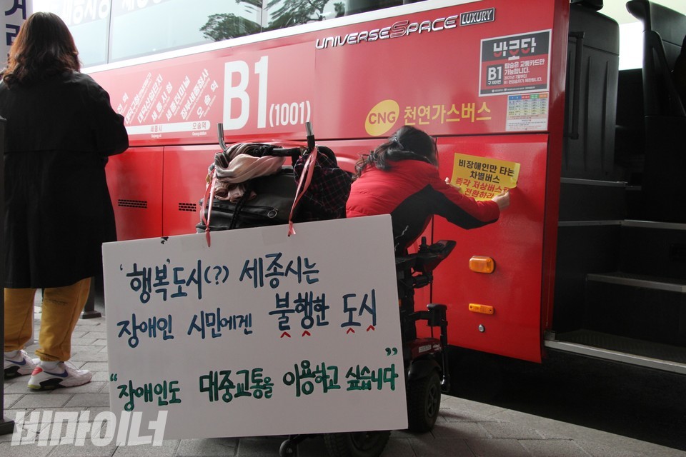 한 활동가가 B1 버스에 ‘비장애인만 타는 차별버스 즉각 저상버스로 전환하라’라고 적힌 피켓을 붙이고 있다. 휠체어에는 ‘행복도시(?) 세종시는 장애인 시민에겐 불행한 도시, 장애인도 대중교통을 이용하고 싶습니다’라고 적힌 피켓을 걸었다. 사진 하민지