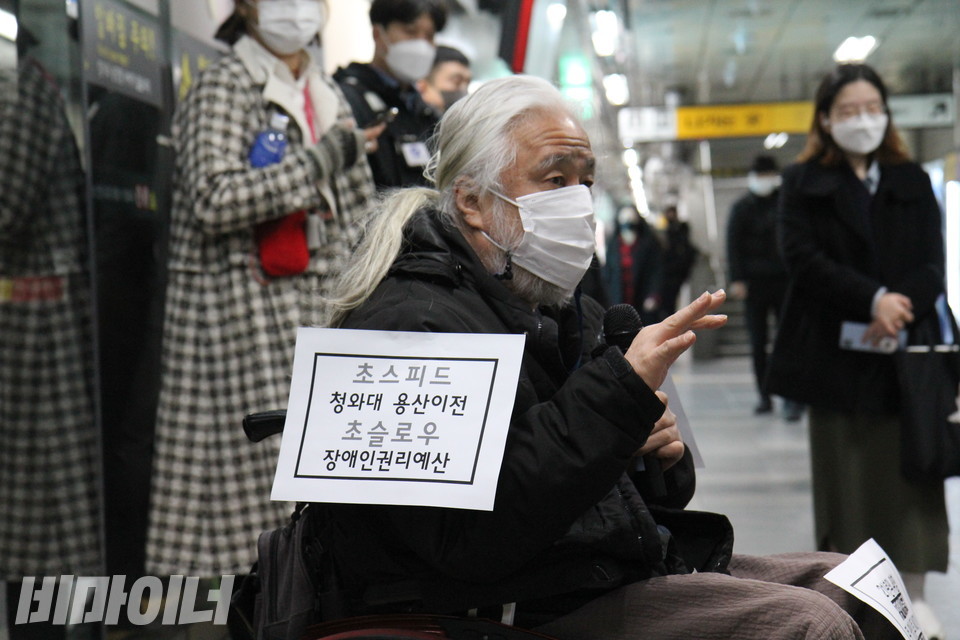 인수위로 향하기 전, 혜화역 승강장 선전전에서 박경석 전장연 상임공동대표가 발언하고 있다. 그의 오른팔에 “초스피드 청와대 용산 이전 / 초슬로 장애인권리예산”이라 적힌 종이가 붙어 있다. 사진 이슬하