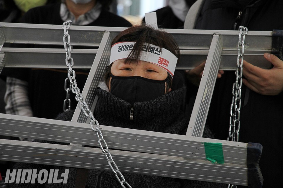 차한선 회원의 목에 쇠사슬과 사다리가 걸려 있다. 그는 이마에 ‘장애인권리예산 쟁취!’라고 적힌 띠를 둘렀다. 사진 하민지