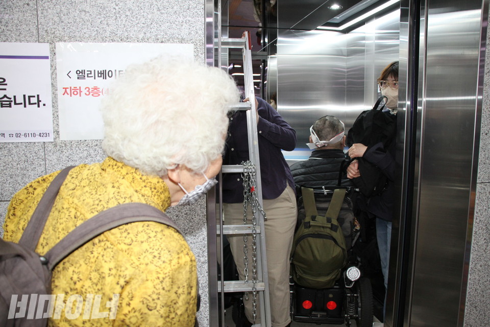 머리가 새하얀 어르신이 엘리베이터에 탑승한 사람들을 바라보고 있다. 엘리베이터 안에는 휠체어 탄 장애인과 비장애인, 사다리 등이 있다. 사진 강혜민