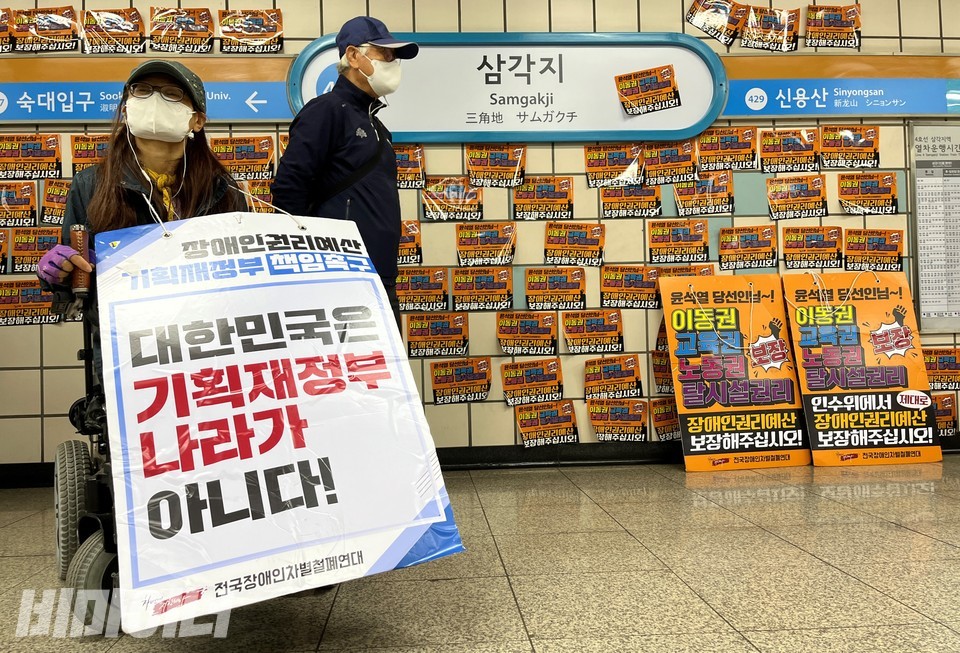 전장연은 오늘(6일)부터, 10일 새 정부 출범에 맞춰 대통령직인수위원회가 있던 3호선 경복궁역에서 4호선 삼각지역으로 투쟁 거점을 변경했다. 뒤로 선전물이 빼곡히 붙어 있다. 한 활동가가 '대한민국은 기획재정부 나라가 아니다'라고 쓰여 있는 팻말을 목에 걸고 있다. 사진 허현덕