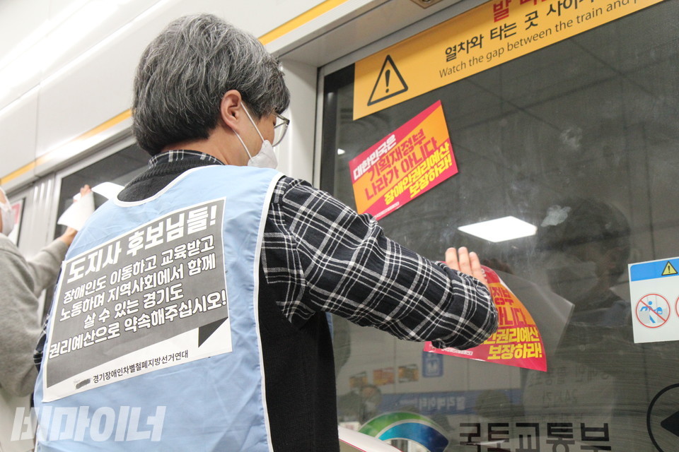 한 활동가가 지하철 스크린도어에 장애인권리예산 보장을 요구하는 스티커를 붙이고 있다. 그는 “도지사 후보님들! 장애인도 이동하고 교육받고 노동하며 지역사회에서 함께 살 수 있는 경기도, 권리예산으로 약속해주십시오!”라고 적힌 조끼를 입고 있다. 사진 이슬하