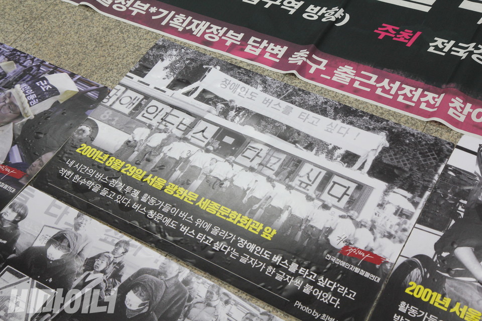바닥에 사진 피켓들이 놓여 있다. 한 피켓에 “2001년 8월 29일 서울 광화문 세종문화회관 앞. 네 시간의 버스 점거 투쟁. 활동가들이 버스 위에 올라가 ‘장애인도 버스를 타고 싶다’라고 적힌 현수막을 들고 있다. 버스 창문에도 ‘버스 타고 싶다’는 글자가 한 글자씩 붙어 있다”고 적혀 있다. 사진 이슬하