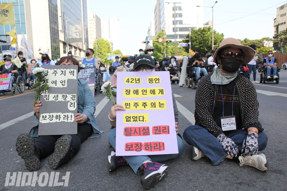활동가들이 도로에 앉아 피켓을 들고 있다.  “42년 동안 장애인에게 민주주의는 없었다. 탈시설권리 보장하라!”,  “광주지역 장애인 자립생활권리 보장하라!”고 적혀 있다. 사진 이슬하