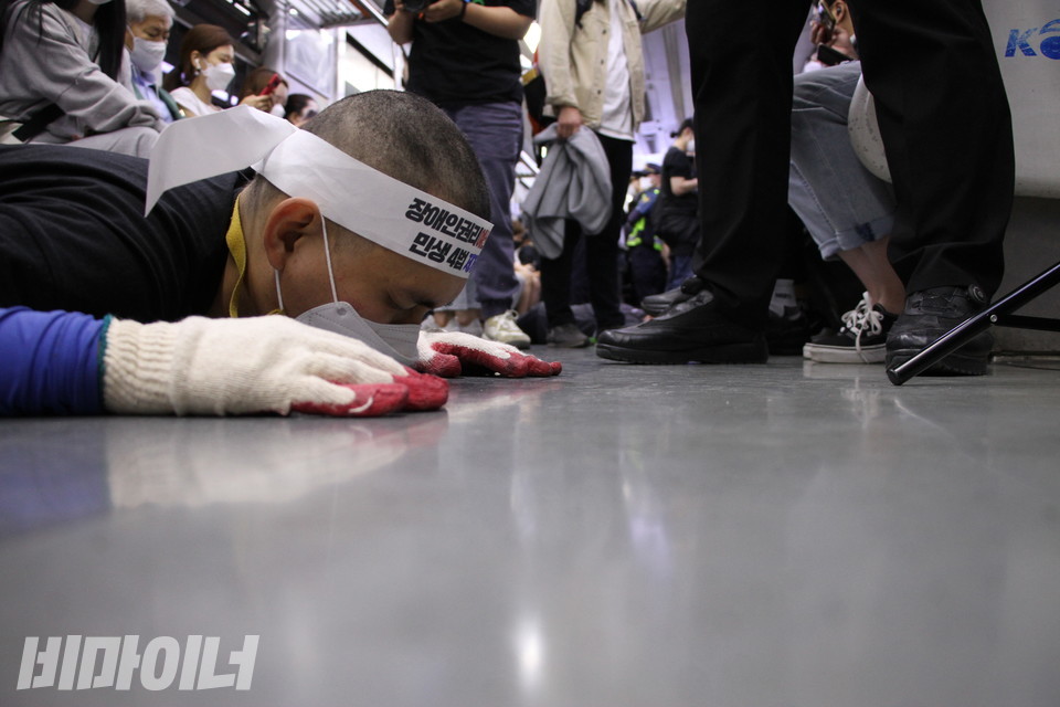 김민석 진주장애인자립생활센터 소장이 지하철 바닥에 힘겨운 듯 고개를 대고 있따. 그 뒤로는 시민들이 의자에 앉아 있다. 서 있는 시민의 신발도 보인다. 사진 허현덕