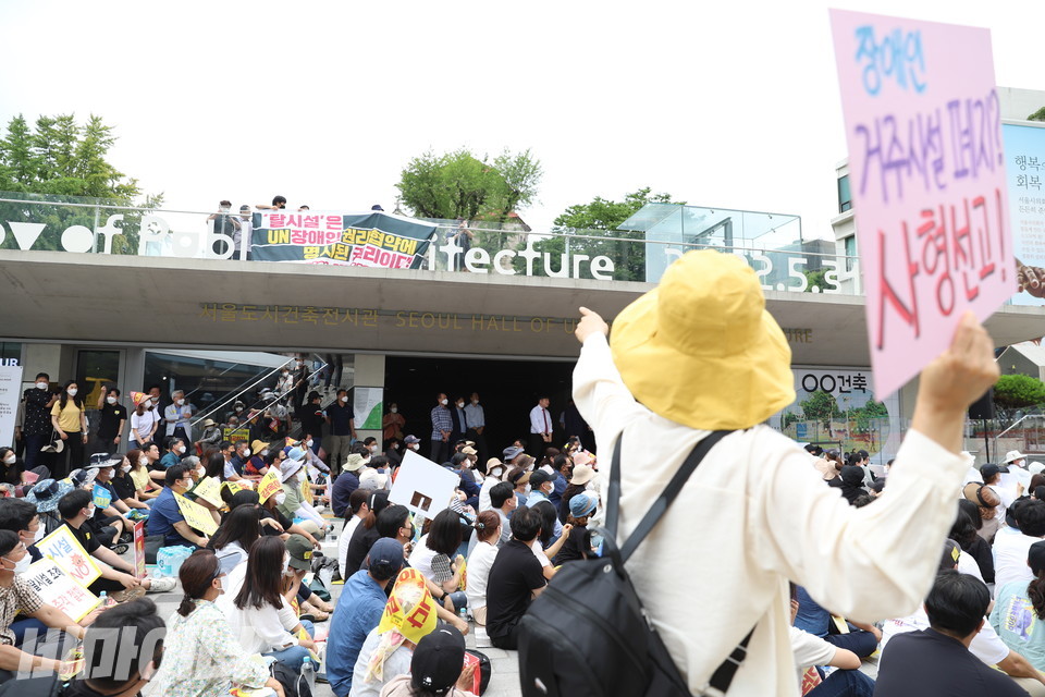 서울시협의회 소속 활동가들이 서울시시설협회 집회가 진행되는 서울도시건축전시관 위에서 “탈시설은 유엔장애인권리협약에 명시된 권리다. 서울시와 서울시의회는 장애인탈시설지원조례 즉각 제정하라”고 적힌 대형 현수막을 내렸다. 이에 대해 탈시설 반대 집회를 열던 사람들이 일제히 고개를 돌려 쳐다보고 있다. “장애인 거주시설 폐지? 사형선고!”라는 피켓을 든 사람이 현수막을 손가락으로 가리키며 소리치고 있다. 사진 강혜민