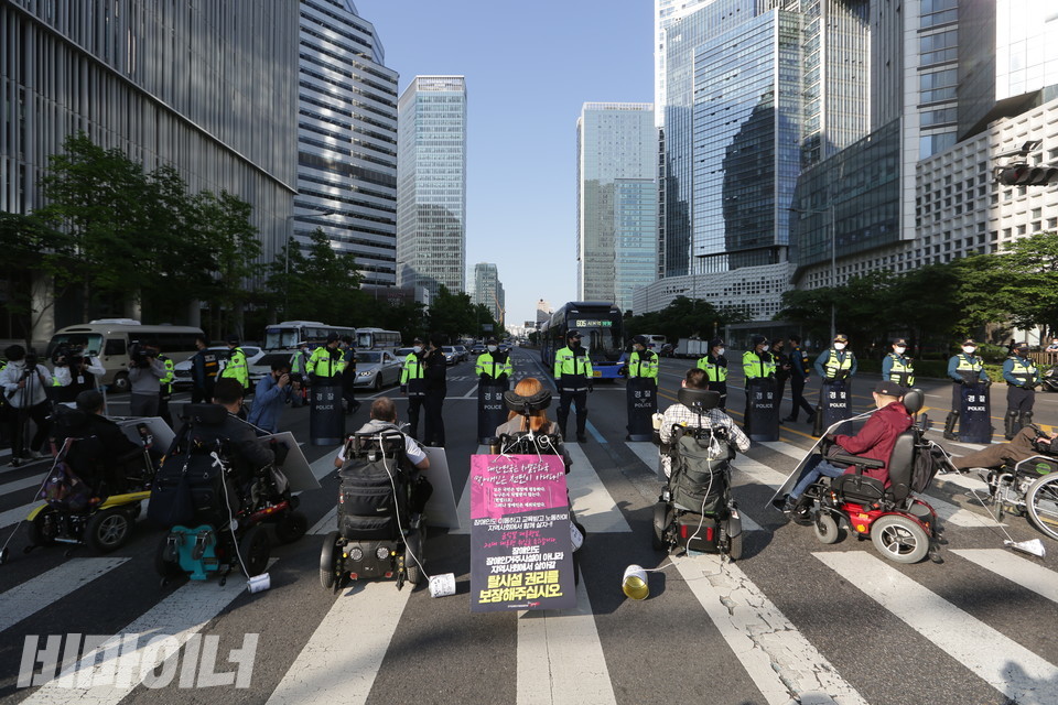 휠체어에 탄 장애인 활동가들이 장애인권리예산을 요구하며 용산구 한강대로를 막아섰다. 맞은편에 방패를 든 경찰이 서 있고, 그 너머로 차들이 보인다. 높은 빌딩숲으로 하늘은 가리어져 있다. 사진 이슬하