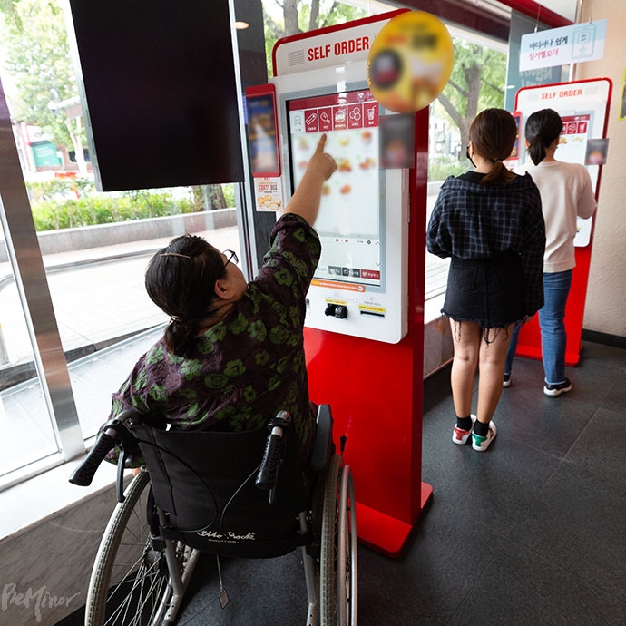 휠체어 이용 장애인이 패스트푸드점에서 무인정보단말기를 이용하려고 손을 뻗고 있다. 사진 비마이너DB