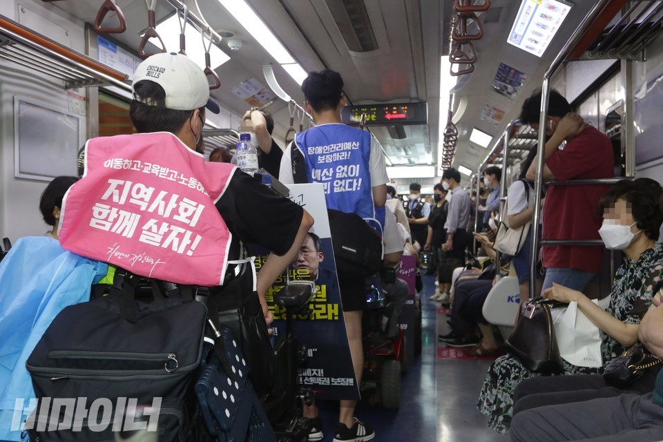 휠체어를 이용하는 활동가들이 일렬로 지하철에 타 있다. “지역사회 함께 살자!”, “예산 없이 권리 없다!”라고 쓰인 조끼를 입고 있다. 사진 이슬하
