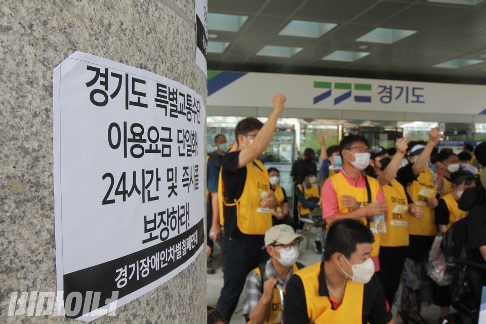 사진 왼편에 보이는 청사 기둥에 “경기도 특별교통수단 이용요금 단일화! 24시간 및 즉시콜 보장하라!”고 적힌 피켓이 붙어 있다. 그 오른편으로 구호를 외치고 있는 기자회견 참가자들이 보인다. 사진 이슬하 