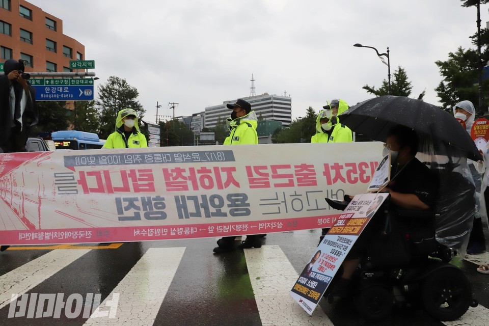 이형숙 서울시장애인자립생활센터협의회 회장이 윤 대통령에게 전달할 공문을 든 채 횡단보도를 건너고 있다. 그의 옆으로 “8월 17일 윤석열 대통령 취임 100일 맞이 제35차 출근길 지하철 탑니다를 예고하는 용와대 행진”이라고 적힌 현수막이 지나가고 있고, 멀리 대통령실 청사가 보인다. 사진 이슬하