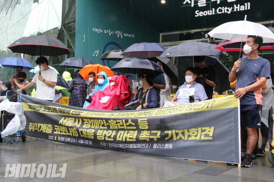 우산을 쓰고 우비를 입은 기자회견 참가자들 앞에 “복지부의 코로나19 재유행 인정, 지자체는 왜 아무런 대책도 마련하지 않는가? 서울시 장애인·홈리스 등 취약계층 코로나19 대응 방안 마련 촉구 기자회견”이라고 적힌 현수막이 놓여있다. 사진 이슬하