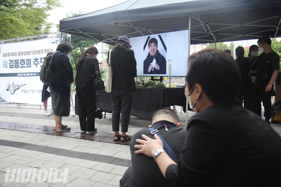 세 사람이 김동호 활동가의 사진 앞에 꽃을 내려놓은 뒤, 고개를 숙인 채 눈을 감고 조용히 그를 생각하고 있다. 멀찌감치 주저앉아 울고 있는 한 사람과 그의 등을 토닥이는 다른 사람이 보인다. 사진 이슬하 