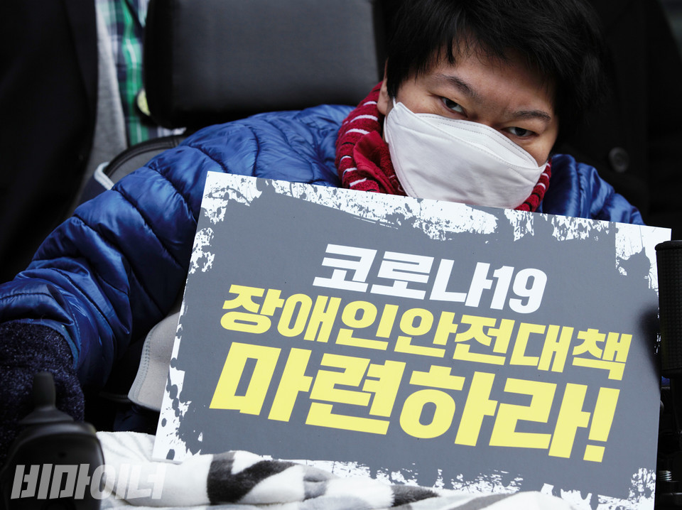 장애인 활동가가 하얀 마스크를 쓰고 “코로나19 장애인 안전대책 마련하라”라고 적힌 손팻말을 들고 있다. 사진 비마이너 DB