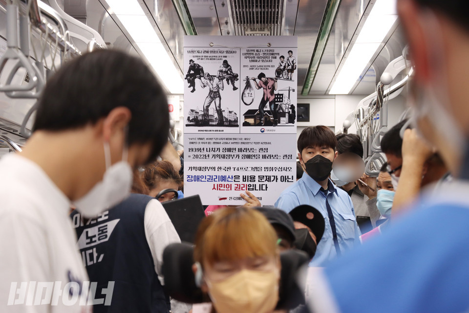 지하철을 탄 장애인 활동가들 뒤로 피켓이 보인다. 피켓에는 “1939년 나치가 장애인 바라보는 관점=2022년 기획재정부가 장애인을 바라보는 관점. 기획재정부의 한국판 T4 프로그램을 멈춰 주십시오. 장애인권리예산은 비용 문제가 아닌 시민의 권리입니다.”라고 적혀 있다. 사진 강혜민