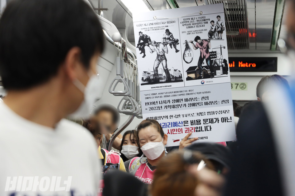 지하철을 탄 활동가들 뒤로 피켓이 보인다. 피켓에는 “1939년 나치가 장애인 바라보는 관점=2022년 기획재정부가 장애인을 바라보는 관점. 기획재정부의 한국판 T4 프로그램을 멈춰 주십시오. 장애인권리예산은 비용 문제가 아닌 시민의 권리입니다.”라고 적혀 있다. 한 활동가가 피곤한 듯 두 눈을 감고 있다. 사진 강혜민