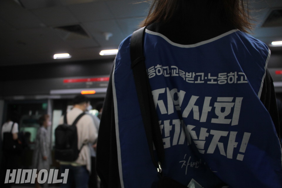 9호선 여의도역 승강장. 한 활동가의 뒷모습이 보인다. 그가 입은 조끼에는 “이동하고, 교육받고, 노동하고, 지역사회 함께 살자!”라고 적혀있다. 그의 앞으로 지하철에 오르는 사람들이 흐릿하게 보인다. 사진 이슬하 