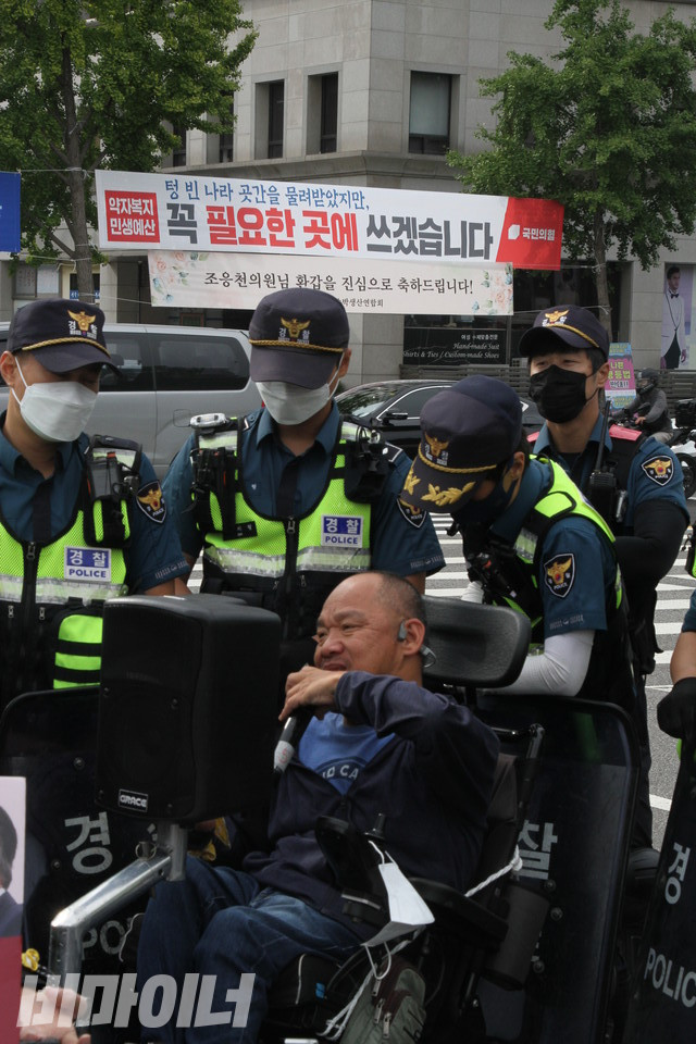 이규식 서울장애인차별철폐연대 대표가 경찰에 둘러싸인 채 발언하고 있다. 그의 뒤로 보이는 횡단보도 위에 “약자복지 민생예산 / 텅 빈 나라 곳간을 물려받았지만, 꼭 필요한 곳에 쓰겠습니다”라고 적힌 국민의힘의 현수막이 걸려있다. 사진 이슬하