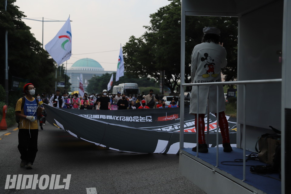 해질녘 행진하는 전장연 활동가들 뒤로 국회의사당 지붕이 보인다. 민중엔터테이너 야마가타트윅스터가 행진 중 공연하고 있다. 사진 복건우