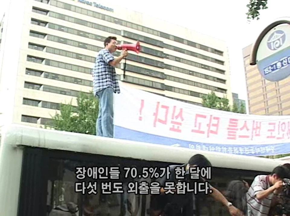 2001년 여름 광화문 세종문화회관 앞 장애인 이동권 투쟁 현장. 서른 살의 문상민 활동가가 버스 위에서 확성기를 들고 “장애인들 70.5%가 한 달에 다섯 번도 외출을 못합니다”라고 외치고 있다. 사진 다큐 ‘버스를 타자’(고 박종필 감독) 캡처