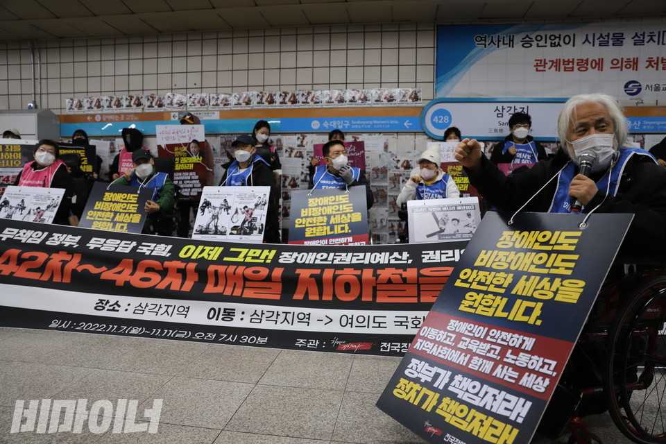 박경석 대표가 ‘장애인도 비장애인도 안전한 세상을 원합니다. 장애인이 안전하게 이동하고 교육받고 노동하고 지역사회에서 함께 사는 세상 정부가 책임져라! 정치가 책임져라!’라고 적힌 피켓을 목에 걸고 ‘투쟁’을 외치고 있다. 박 대표 뒤에 있는 현수막에는 ‘무책임 정부, 무응답 국힘, 이제 그만! 42차~46차 매일 지하철을 탑니다’라고 적혀 있다. 사진 하민지