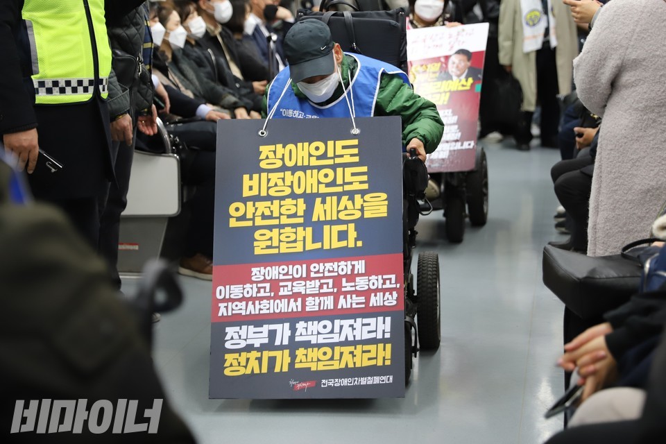 한 장애인 활동가가 ‘장애인도 비장애인도 안전한 세상을 원합니다. 장애인이 안전하게 이동하고 교육받고 노동하고 지역사회에서 함께 사는 세상 정부가 책임져라! 정치가 책임져라!’라고 적힌 피켓을 목에 걸고 지하철에 탔다. 다른 승객은 좌석에 앉아 있다. 사진 하민지