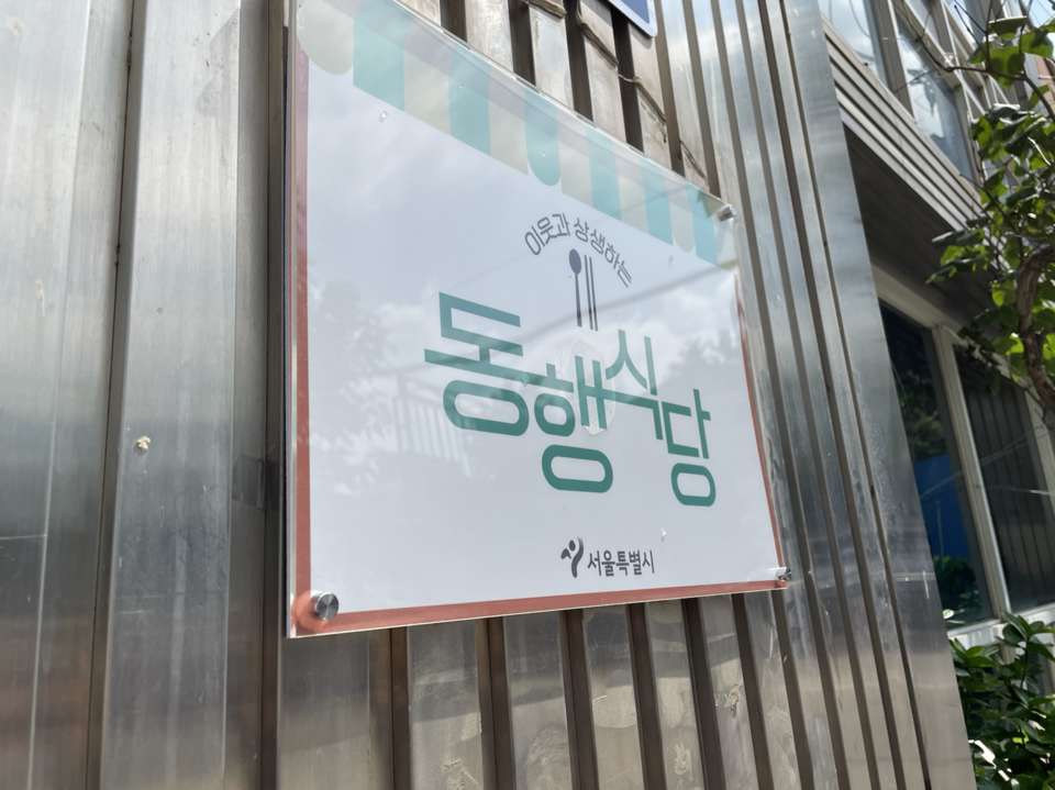 동자동의 한 식당에 붙어 있는 동행식당 표지판. 큰 초록색 글씨로 적힌 ‘동행식당’ 위에 수저 그림이 있고, ‘이웃과 상생하는’이라고 적혀 있다. 그 아래에는 서울특별시 로고가 있다. 사진 홈리스행동