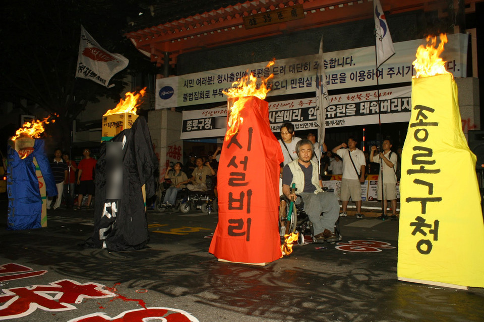 2006년 8월 9일, 종로구청 앞에서 S재단 산하 장애인거주시설에서 발생한 시설 비리, 인권유린을 규탄하며 화형식을 하고 있다. 불을 붙이는 박경석 대표 뒤로 김정하 활동가가 있다. 사진 전진호