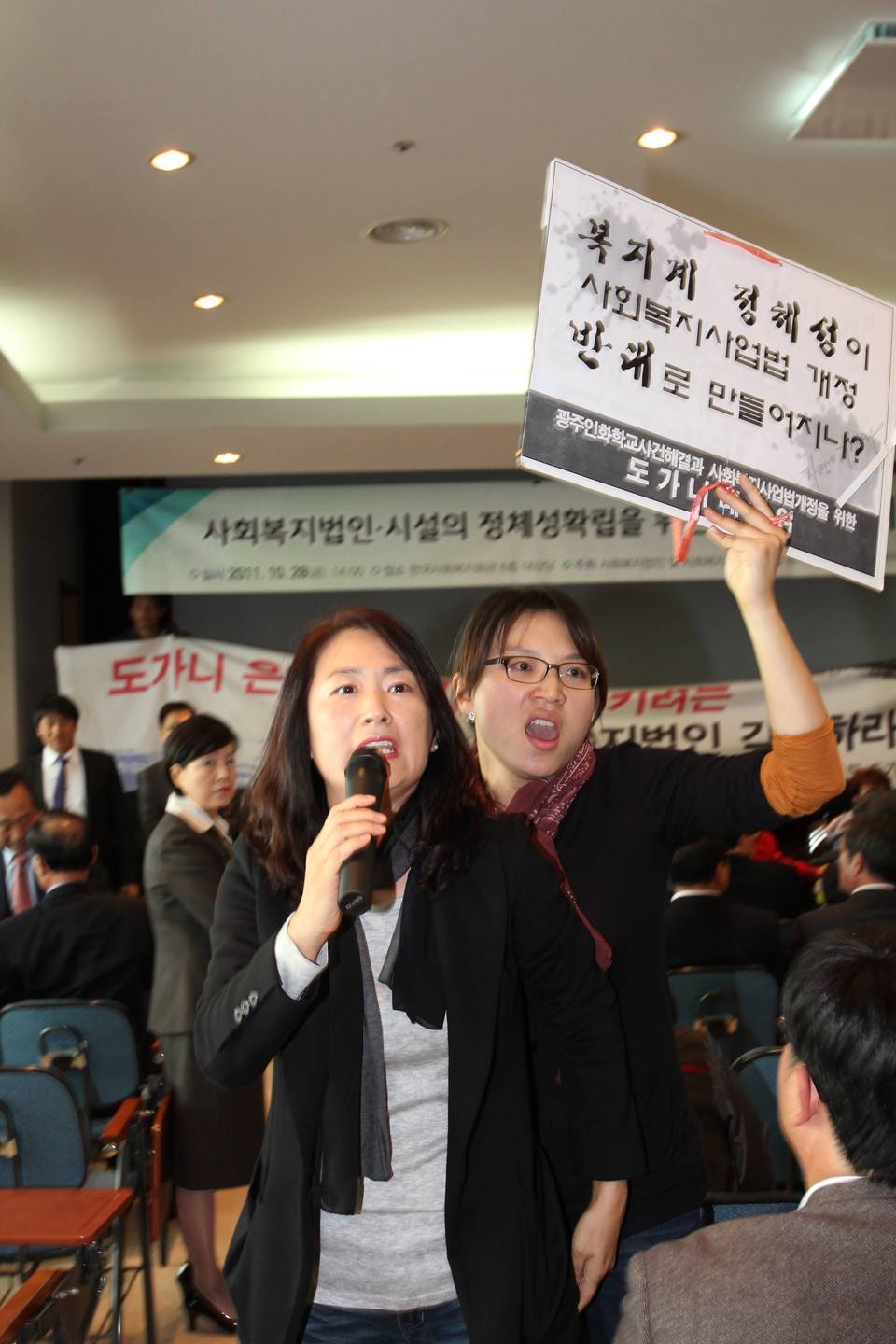 2011년 10월 28일 서울 공덕동 사회복지회관 6층 대회의실에서 ‘사회복지법인·시설의 정체성 확립을 위한 범사회복지 전진대회’가 열렸다. 이날 행사는 공익이사제 도입이 담긴 사회복지사업법 개정안을 반대하는 목소리를 모으기 위한 자리였다. 장애계 활동가들은 이러한 사회복지계의 행보를 규탄하며 기습 시위를 벌였다. 한 시설 원장이 마이크를 잡고 발언하자, 그 뒤에서 김정하 활동가가 “복지계 정체성이 사회복지법 개정 반대로 만들어지나?”라고 적힌 피켓을 들고 규탄하고 있다. 사진 전진호