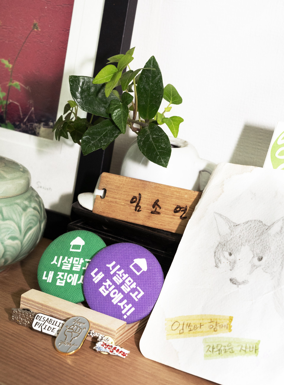‘임소연’ 이름이 새겨진 나무 열쇠고리, ‘시설 말고 내 집에서’라는 문구가 새겨진 배지가 크게 보인다. 서울장애인차별철폐연대, 장애와인권발바닥행동에서 만들어 회원들에게 나눠준 것들이다. 그 옆에는 ‘임쏘(임소연 애칭)와 함께 자유로운 자바’라는 문구와 함께 임소연 활동가와 함께 살았던 고양이 자바 캐리커처 그림이 전시되어 있다. 디스어빌러티 프라이드(DISABILITY PRIDE), 차별에 저항하라 등의 배지도 아기자기하게 놓여 있다. 사진 현다혜 