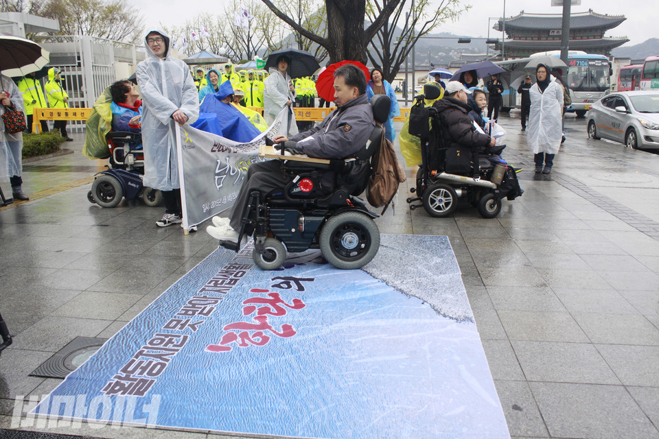 2015년 4월 14일, 한자협은 만 65세가 넘으면 활동지원서비스가 중단되는 현실을 규탄하며 정부서울청사 앞에서 기자회견을 했다. 곧 65세가 다가오는 한 장애인 활동가가 자립생활이 가로막히는 ‘한탄의 강’을 건너는 퍼포먼스를 하고 있다. 오른쪽 뒤에서 임소연 활동가가 이를 지켜보고 있다. 사진 강혜민 