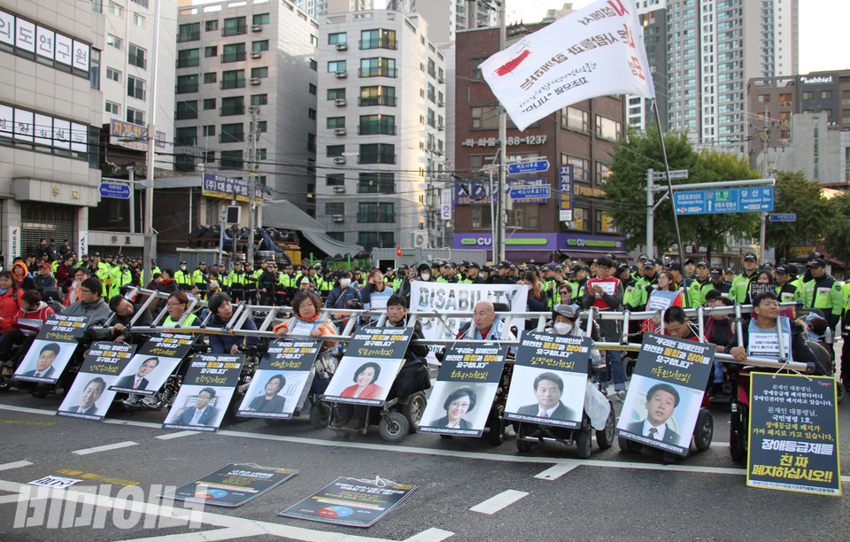 2018년 10월 26일, 한자협 소속 활동가들이 국회에 장애인예산을 촉구하며 목에 사다리와 쇠사슬을 두르고 도로를 점거하고 있다. 그 뒤로는 한자협을 상징하는 디스어빌리티 프라이드(DISABILITY PRIDE, 장애 자부심) 현수막이 펼쳐져 있다. 사진 강혜민 
