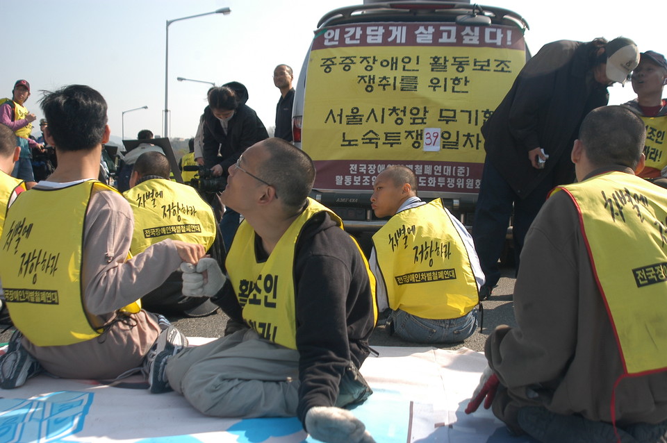 2006년 4월 27일, 장애인 활동가들이 활동지원제도화를 촉구하며 한강대교를 점거했다. 사람들이 “차별에 저항하라. 전국장애인차별철폐연대”라고 적힌 몸피씨를 입고서 휠체어에서 내려와 아스팔트 바닥에 앉아 있다. 사진 n_m