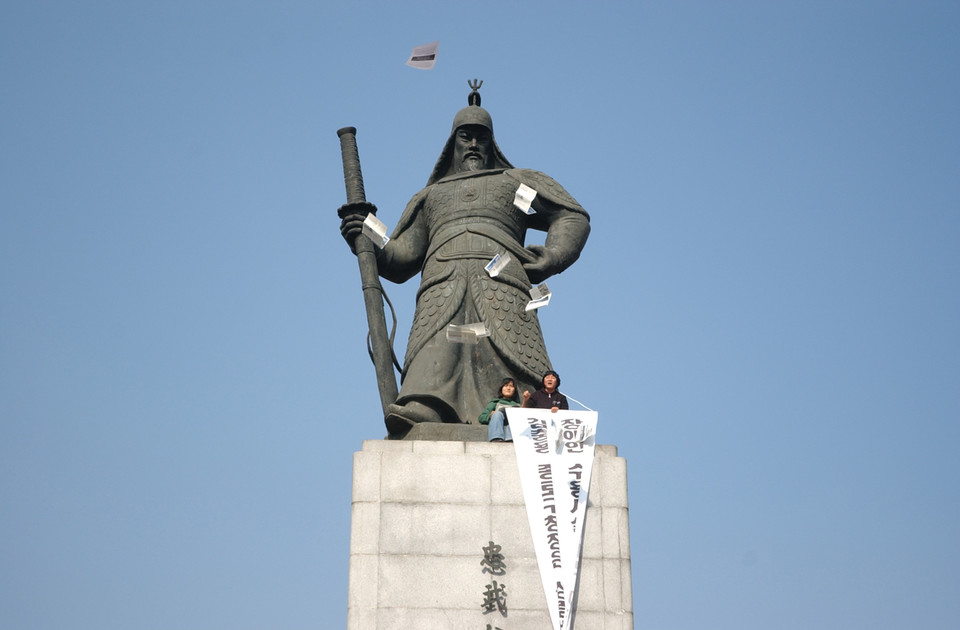 이순신 동상 위에서 현수막을 붙잡고 선전물을 뿌리는 두 활동가. 사진 n_m 