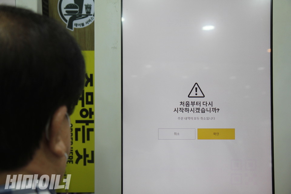 시각장애인 이병길 씨가 무인정보단말기(키오스크)로 음식을 주문하고 있다. 무인정보단말기 화면에는 “(삼각형 안에 느낌표가 그려진 주의 표시와 함께) 처음부터 다시 시작하시겠습니까?”라고 쓰여 있고, 작은 글씨로 “주문 내역이 모두 취소됩니다”라고 적혀 있다. 그 아래 왼쪽에는 취소 버튼이, 오른쪽에는 확인 버튼이 있다. 사진 비마이너DB