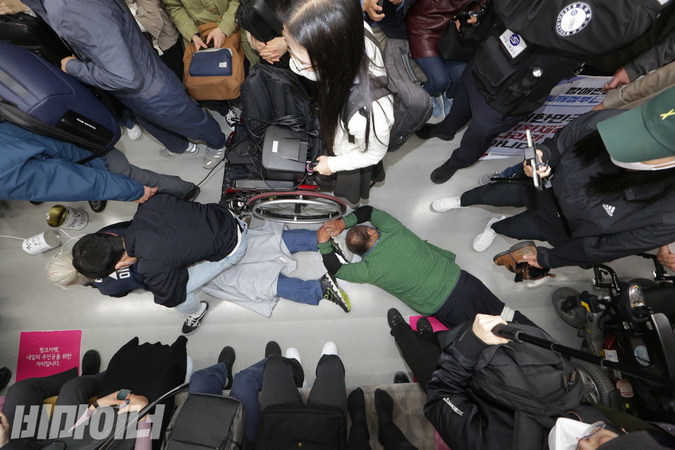 지하철 천장에서 바라본 박경석 대표와 배재현 대의원의 모습. 엎드려 있는 두 사람 주변을 승객과 지하철 보안관 등이 에워싸고 있다. 사진 비마이너DB