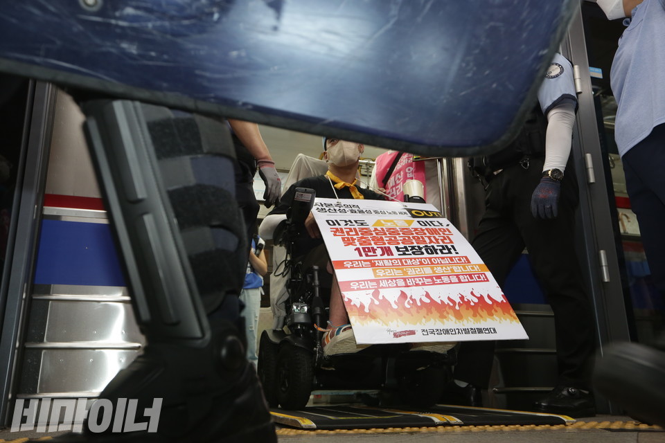 한 장애인 활동가가 “이것도 노동이다. 권리중심중증장애인맞춤형공공일자리 1만개 보장하라”는 문구가 적힌 피켓을 들고 지하철 입구에 서있다. 경찰 방패로 가려져 그의 얼굴은 보이지 않는다. 사진 비마이너DB